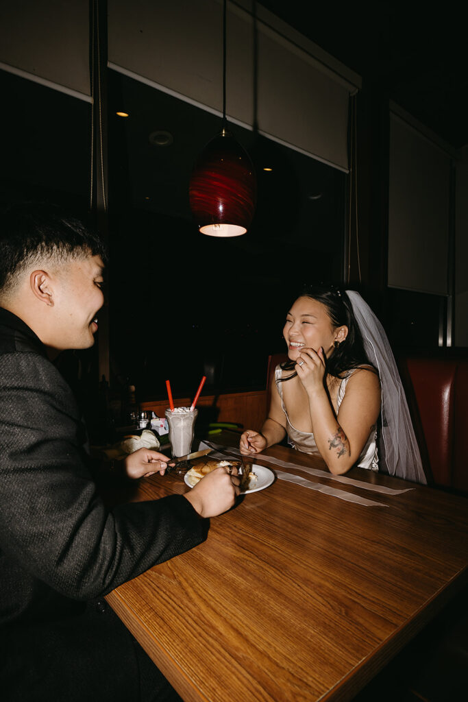 Two people sitting across from each other in a restaurant booth, wearing formal attire and sunglasses, under a red hanging light.