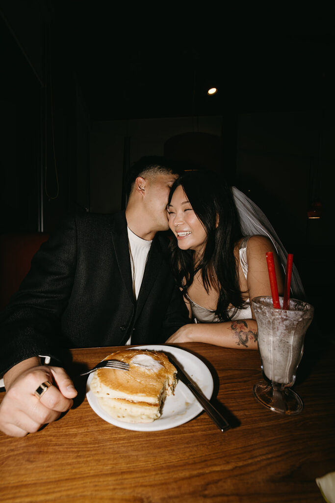 Two people sitting across from each other in a restaurant booth, wearing formal attire and sunglasses, under a red hanging light.