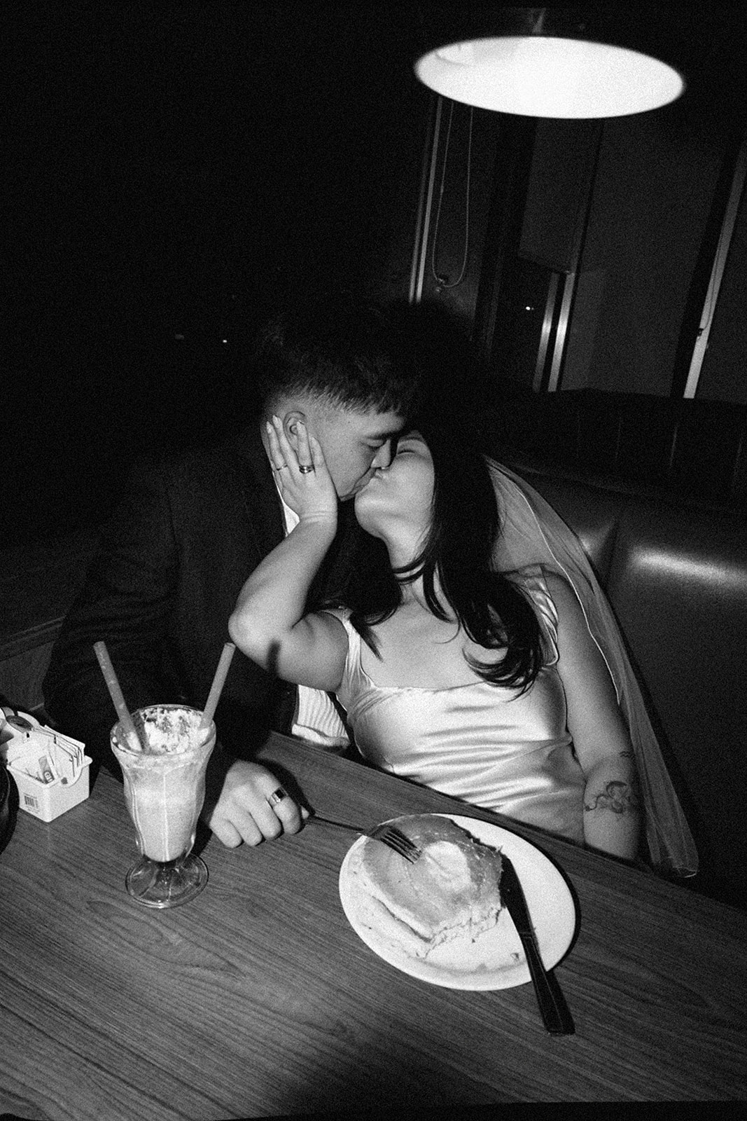 Two people sitting across from each other in a restaurant booth, wearing formal attire and sunglasses, under a red hanging light.