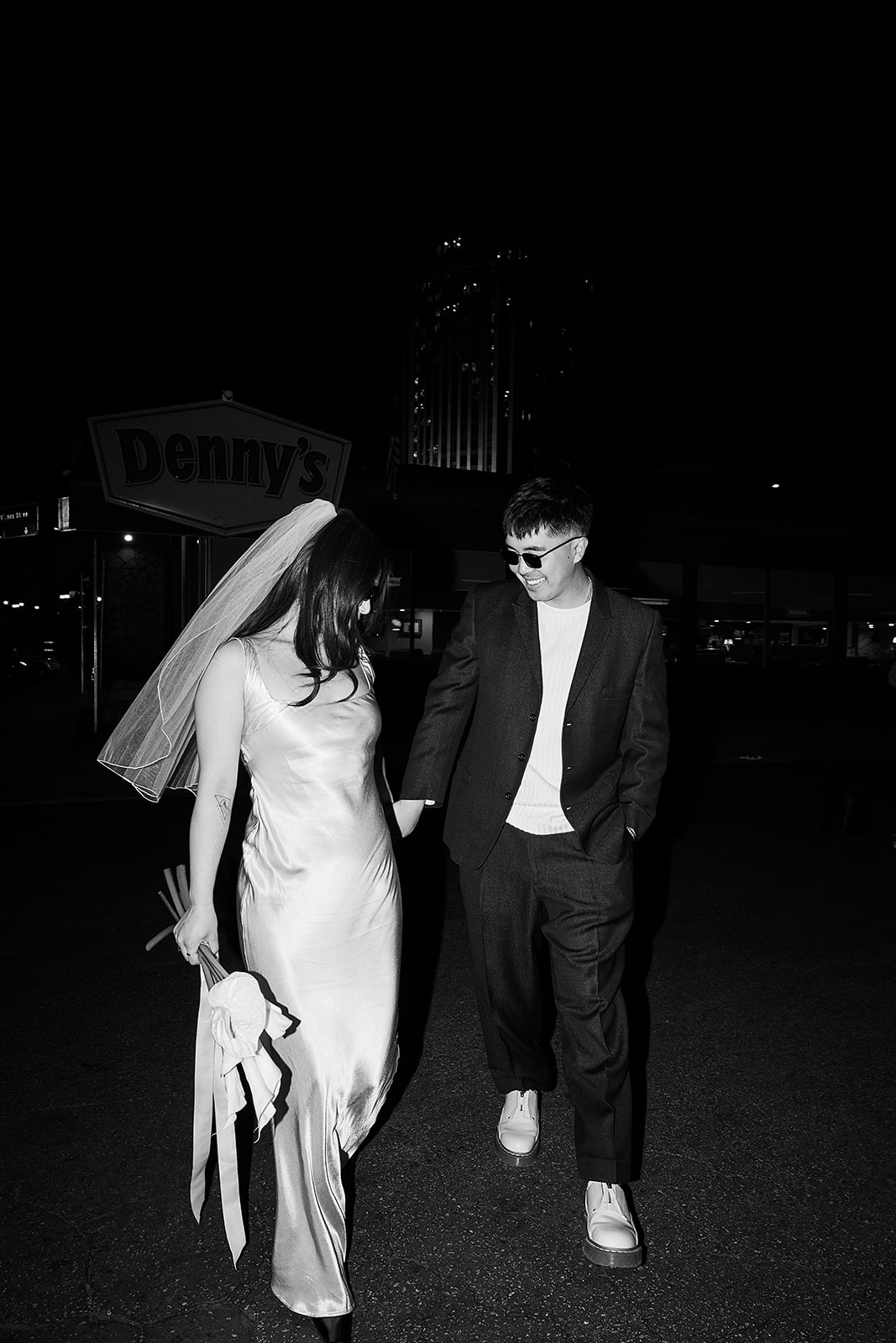 Two people sitting across from each other in a restaurant booth, wearing formal attire and sunglasses, under a red hanging light.