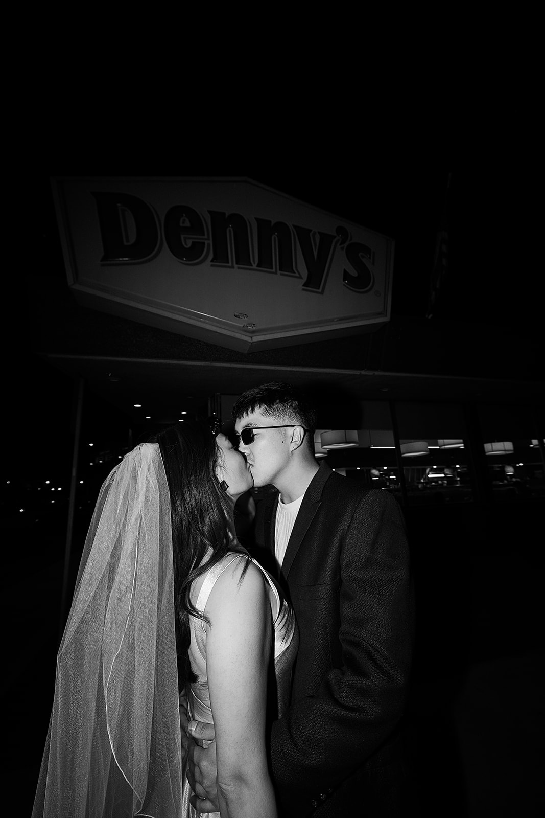 A bride and groom share a kiss in front of a Denny's restaurant at night.