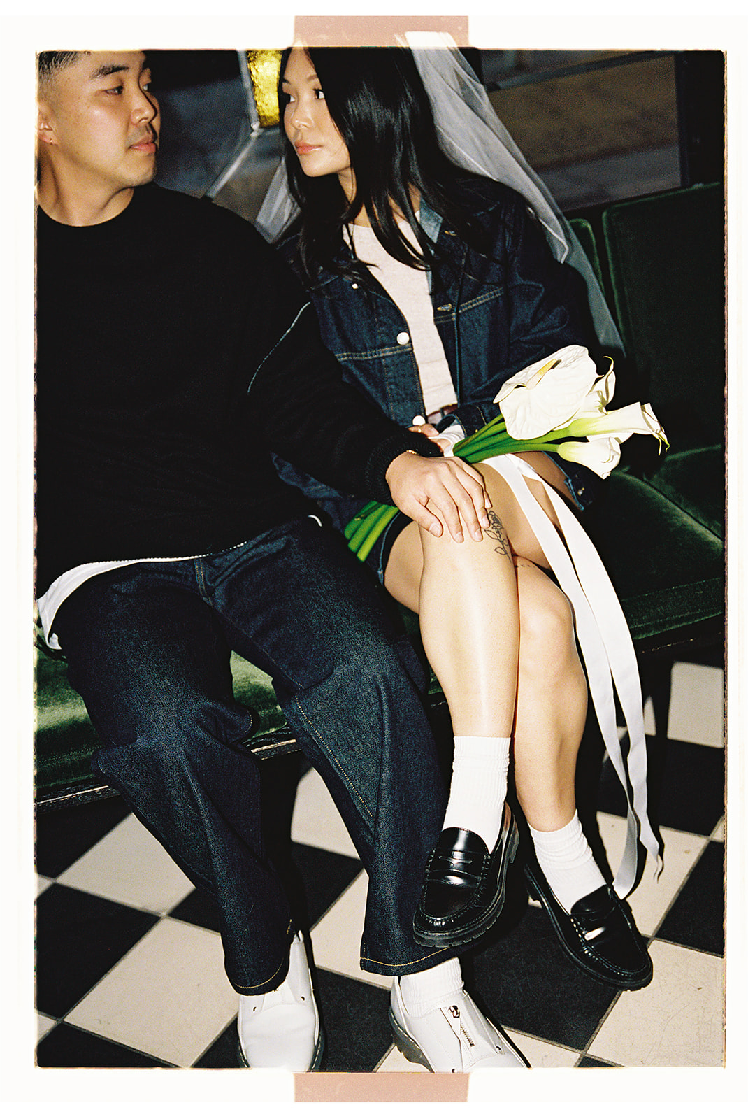 A couple sits closely on a bench; the woman holds a bouquet and wears a veil. They both wear dark outfits and white footwear. The floor has a black and white checkerboard pattern.