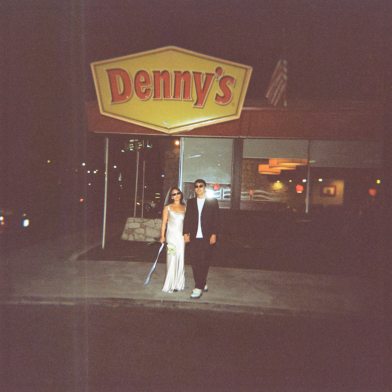 A couple stands outside a Denny's restaurant at night, dressed in formal attire.