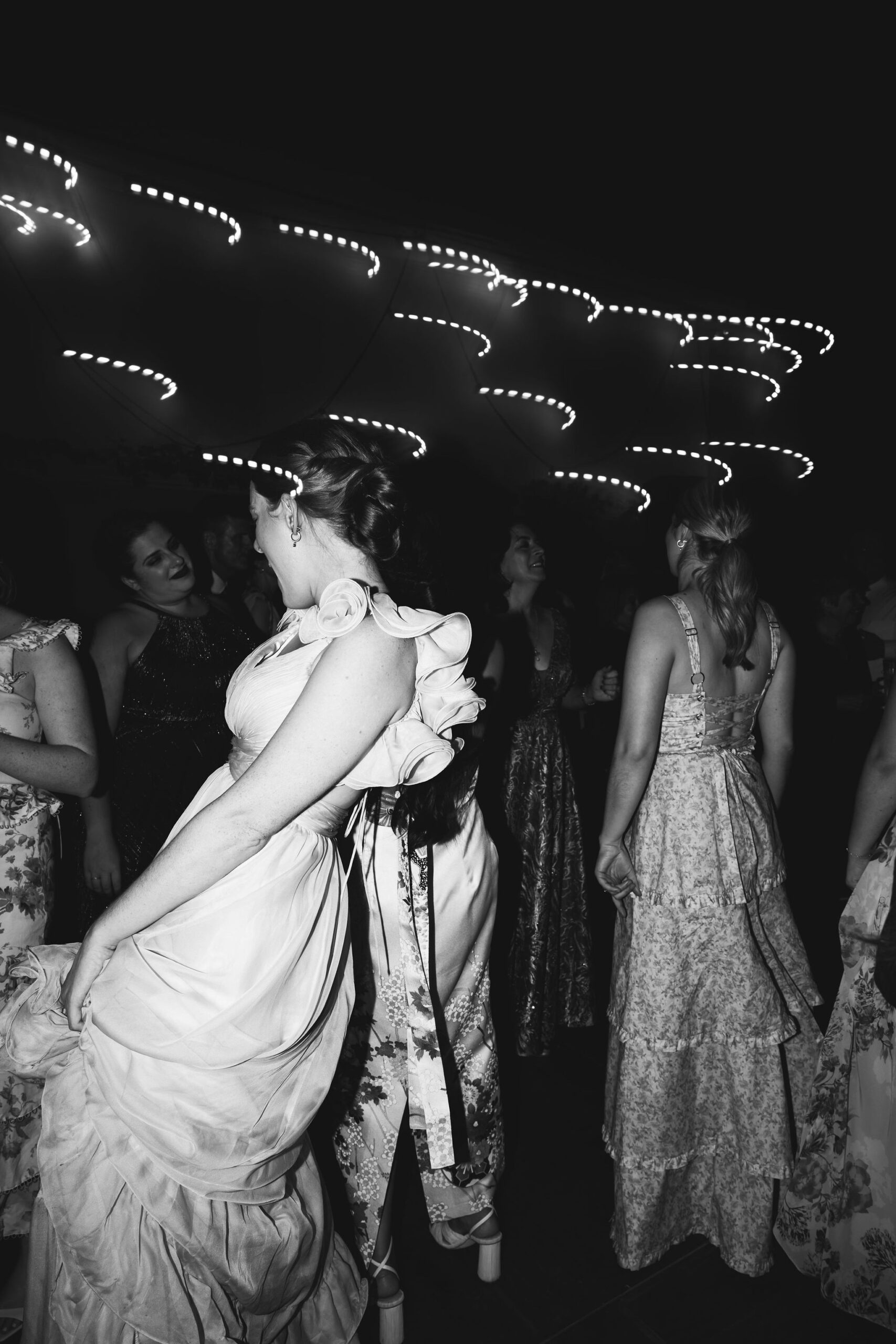 Two women in formal dresses are joyfully dancing and holding drinks at a nighttime event.