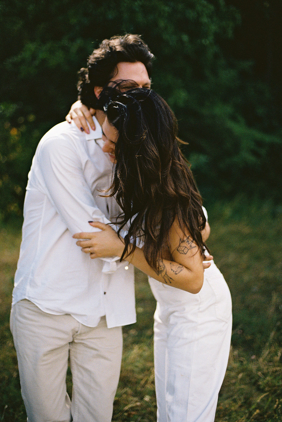 A couple dressed in white embraces outdoors, with greenery in the background during their candid engagement photos