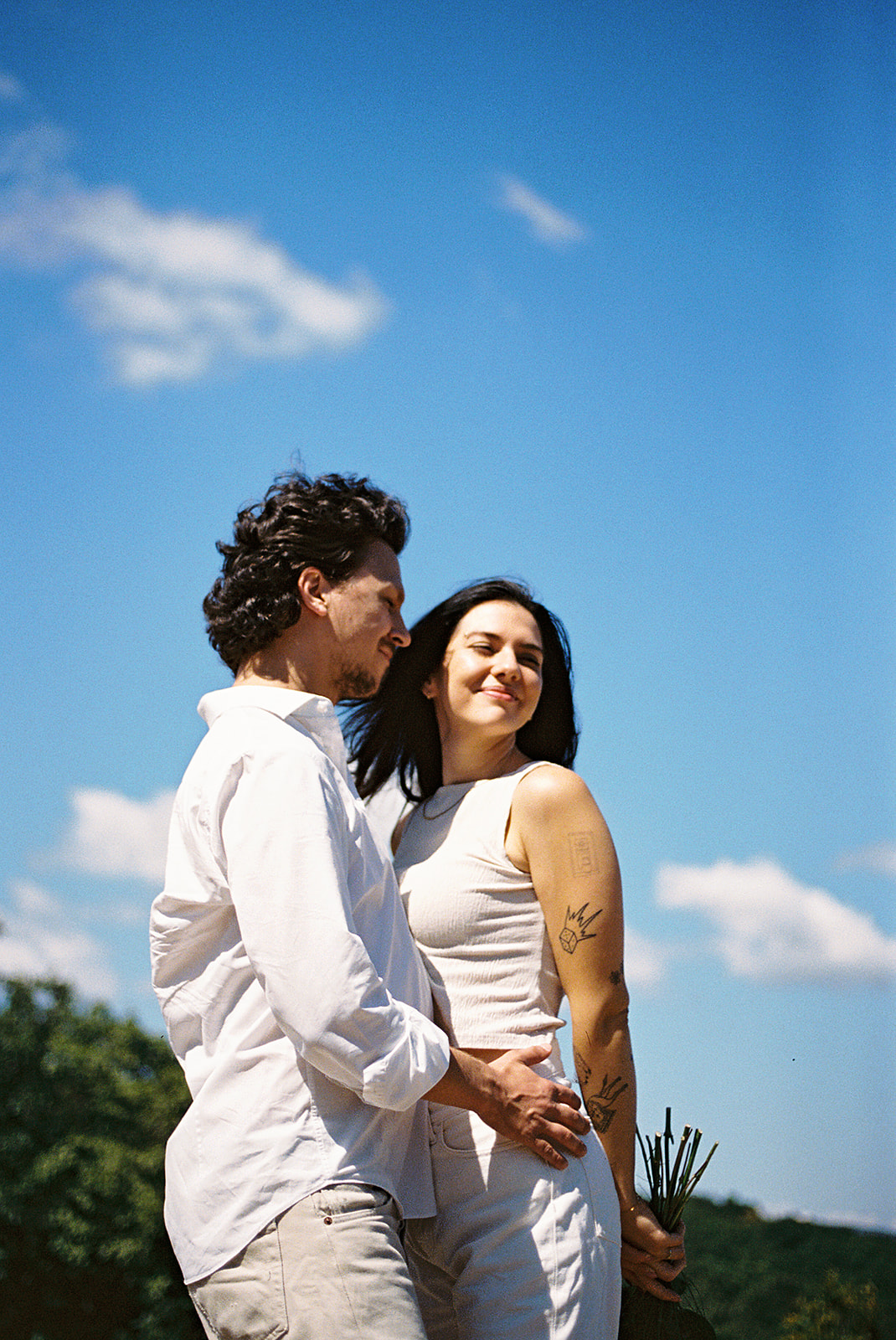 A couple dressed in white embraces outdoors, with greenery in the background during their candid engagement photos