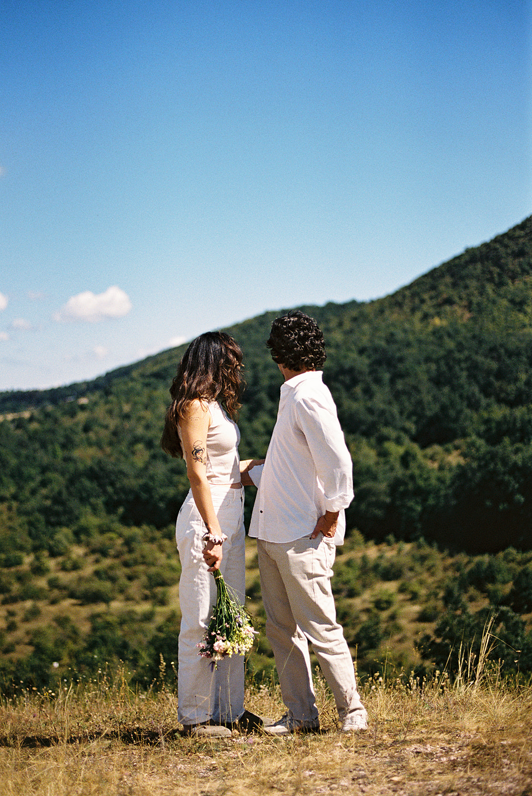 A couple dressed in white embraces outdoors, with greenery in the background during their candid engagement photos