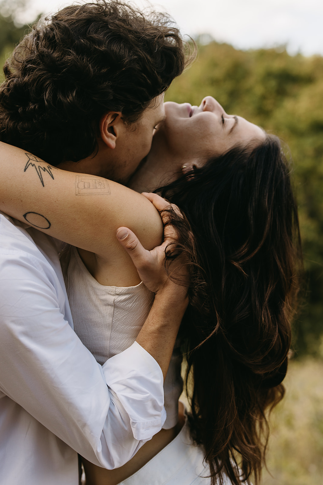 A couple embraces outdoors, the man holding the woman close while she leans back, eyes closed. They are surrounded by greenery.