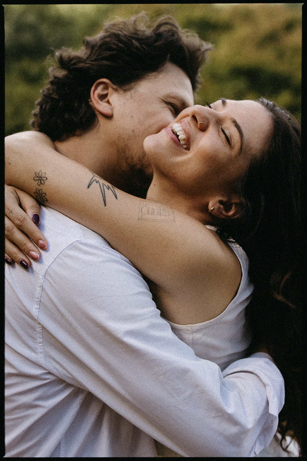 A couple embraces outdoors, smiling and wearing casual attire. The background is blurred greenery.