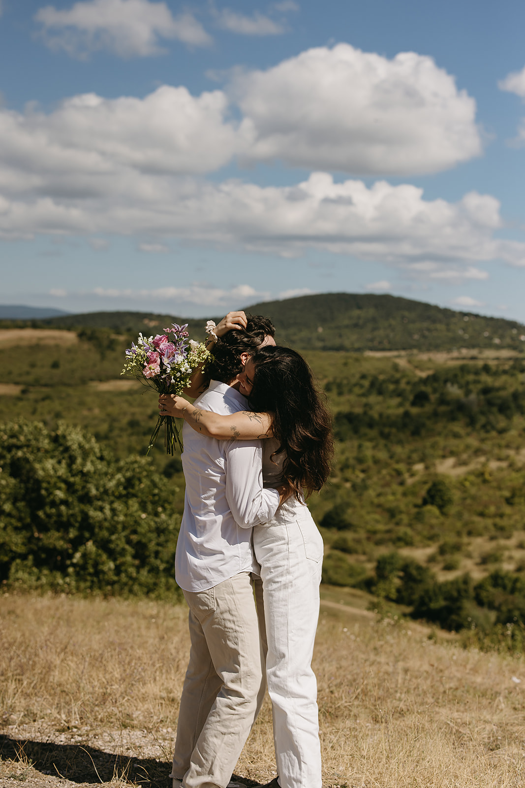 A couple dressed in white embraces outdoors, with greenery in the background during their candid engagement photos