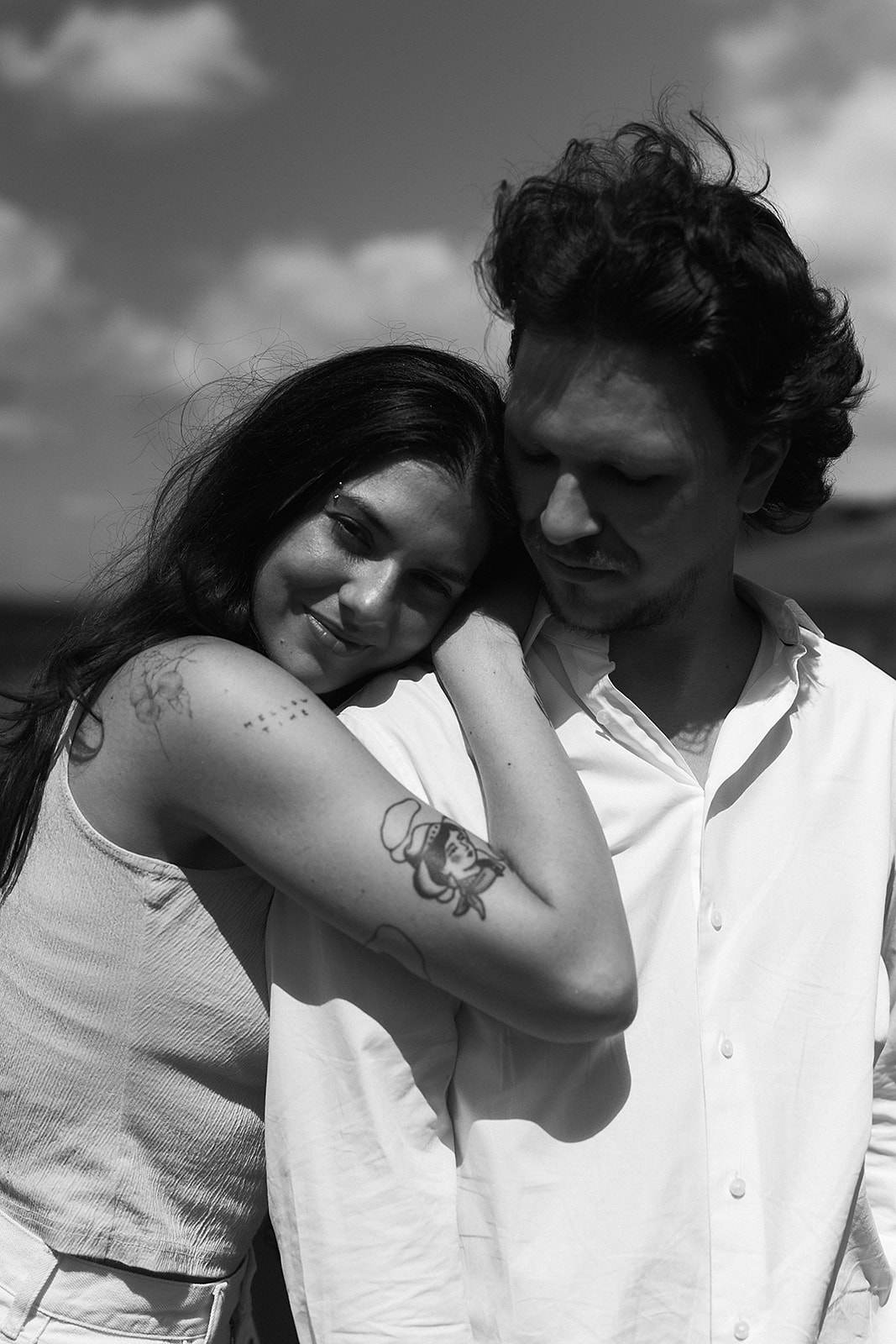 A man and woman stand in an open field under a partly cloudy sky. The woman shields her eyes with her hand and holds flowers. Both wear casual white clothing.