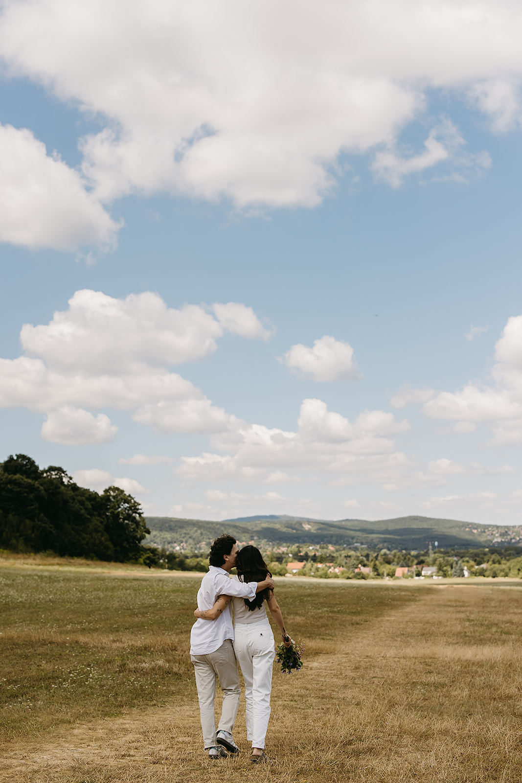 A man and woman stand in an open field under a partly cloudy sky. The woman shields her eyes with her hand and holds flowers. Both wear casual white clothing.