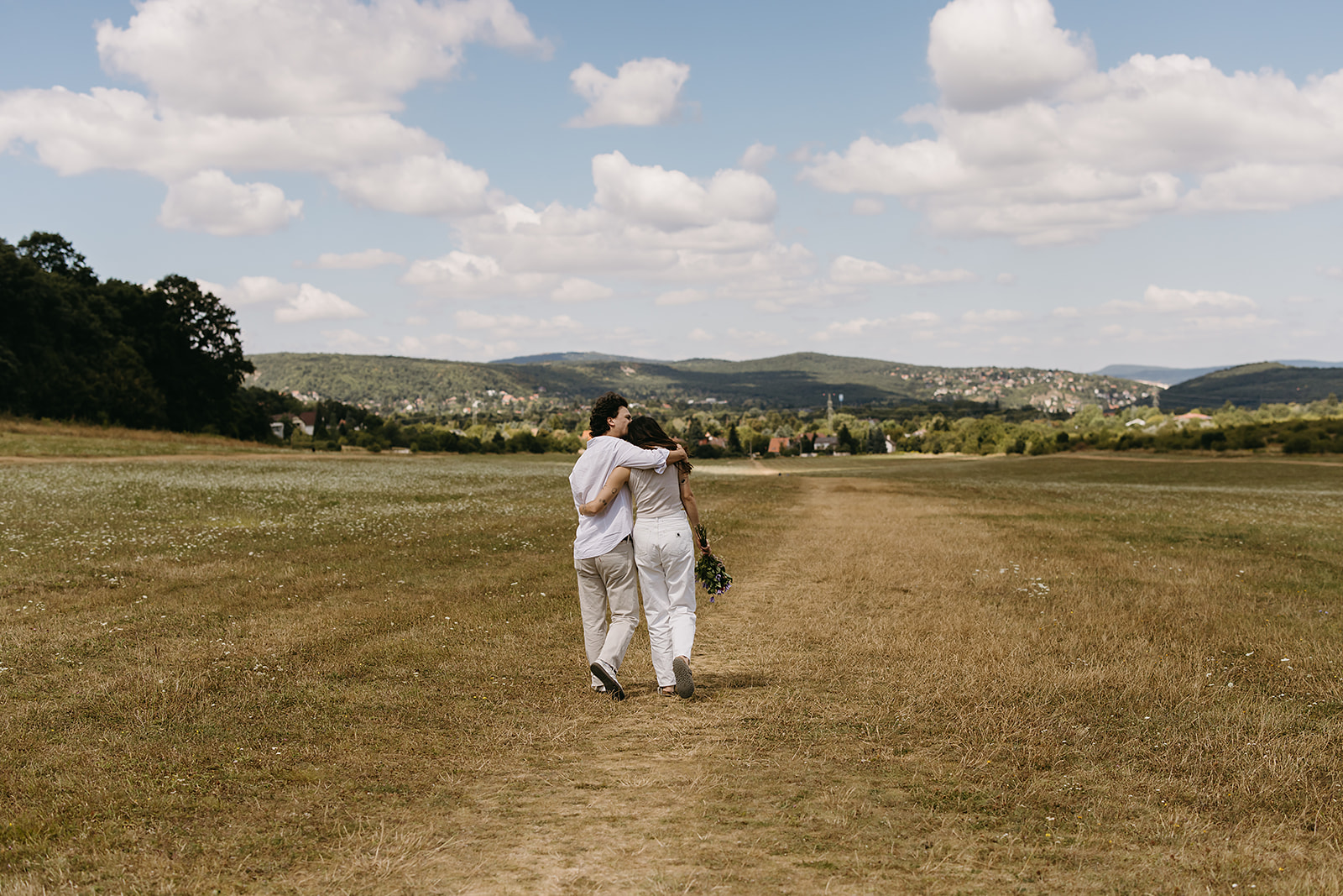 A man and woman stand in an open field under a partly cloudy sky. The woman shields her eyes with her hand and holds flowers. Both wear casual white clothing.