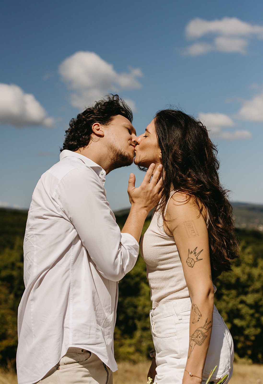 A couple dressed in white embraces outdoors, with greenery in the background during their candid engagement photos