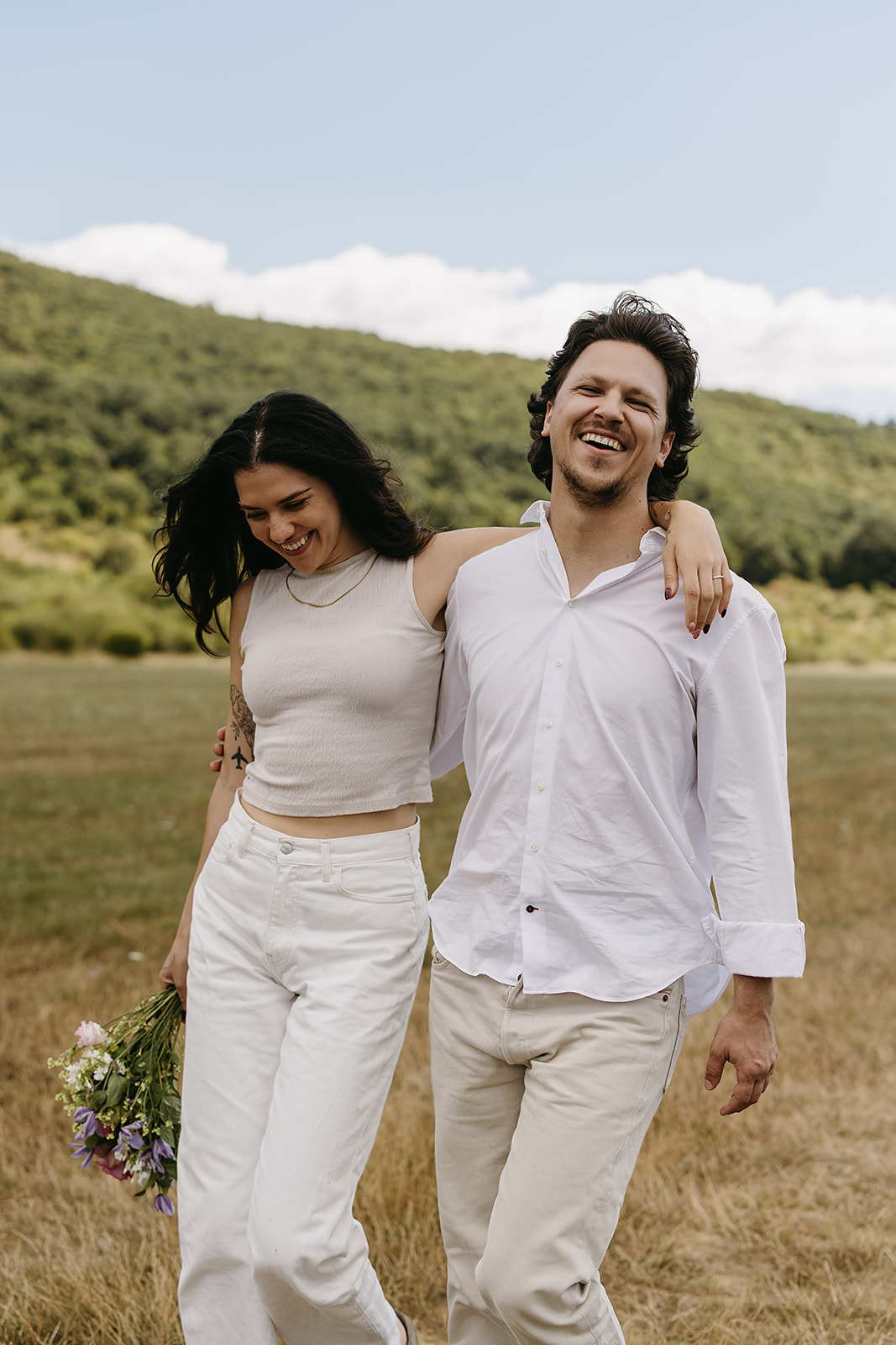 A man and woman stand in an open field under a partly cloudy sky. The woman shields her eyes with her hand and holds flowers. Both wear casual white clothing.