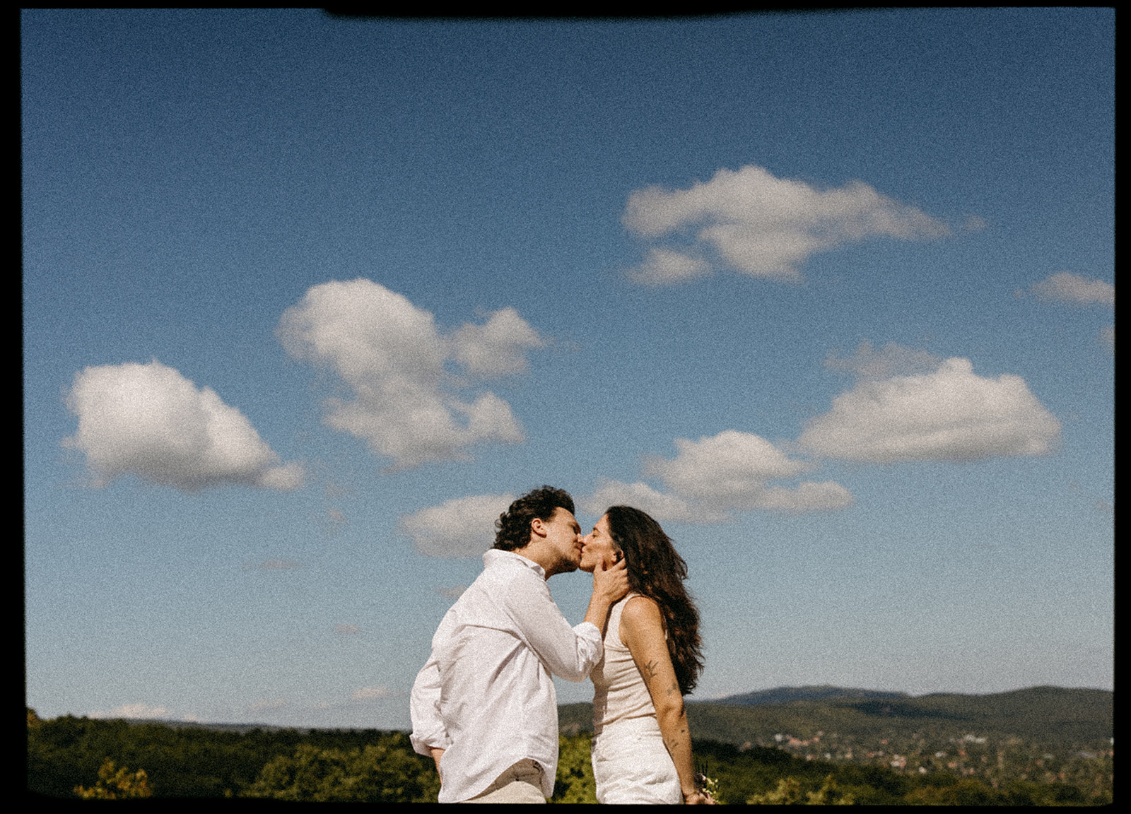 A couple dressed in white embraces outdoors, with greenery in the background during their candid engagement photos