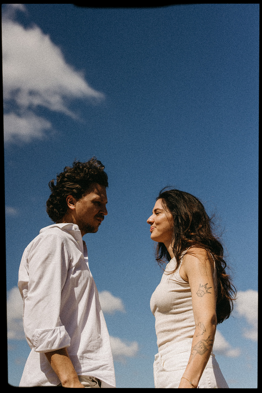 A couple dressed in white embraces outdoors, with greenery in the background during their candid engagement photos