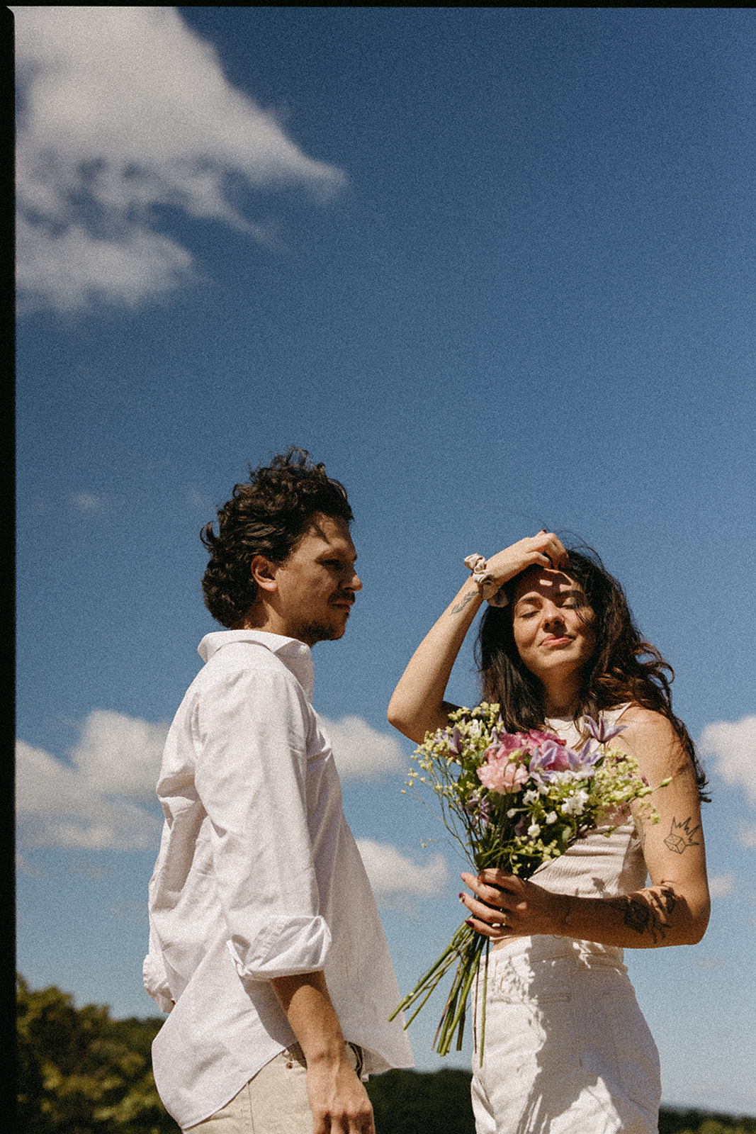 A couple dressed in white embraces outdoors, with greenery in the background during their candid engagement photos