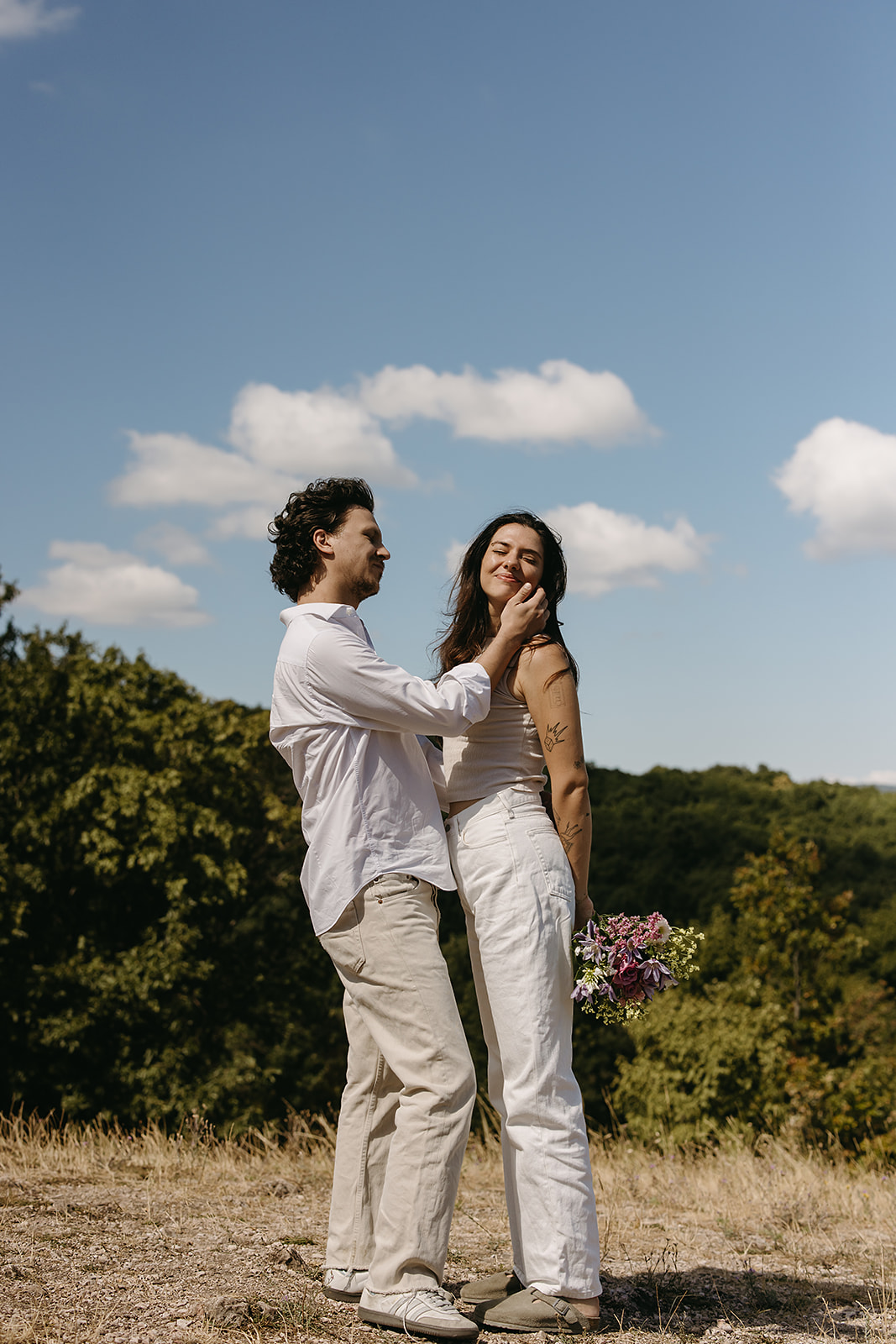 A couple dressed in white embraces outdoors, with greenery in the background during their candid engagement photos