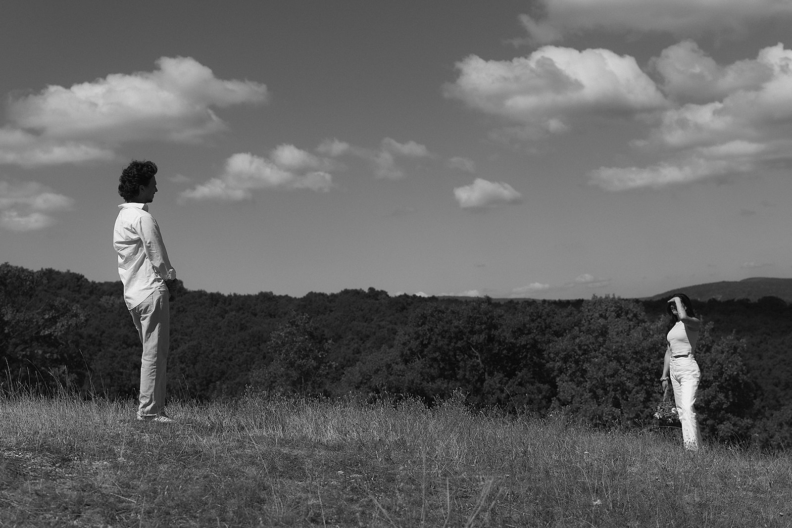 A couple dressed in white embraces outdoors, with greenery in the background during their candid engagement photos