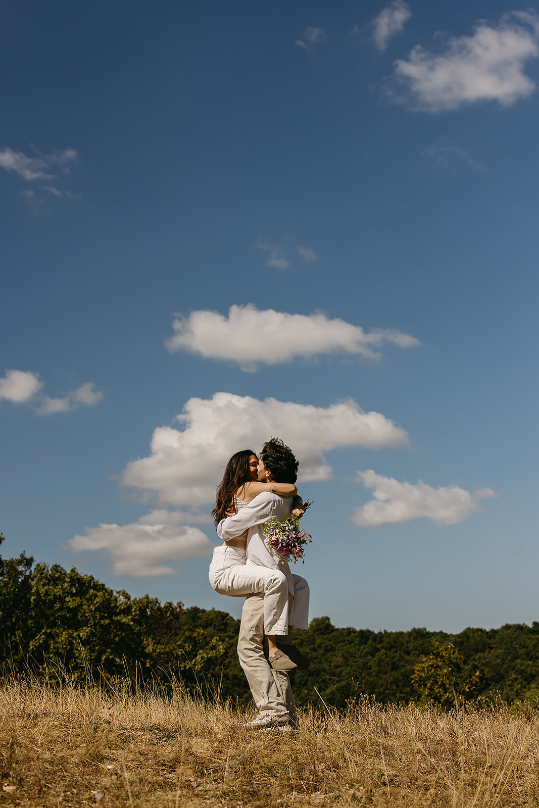 A couple dressed in white embraces outdoors, with greenery in the background during their candid engagement photos