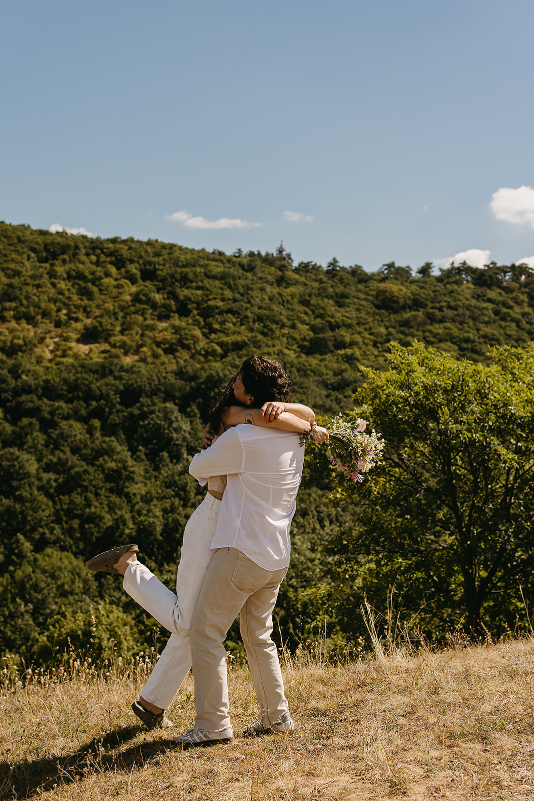 A couple dressed in white embraces outdoors, with greenery in the background during their candid engagement photos