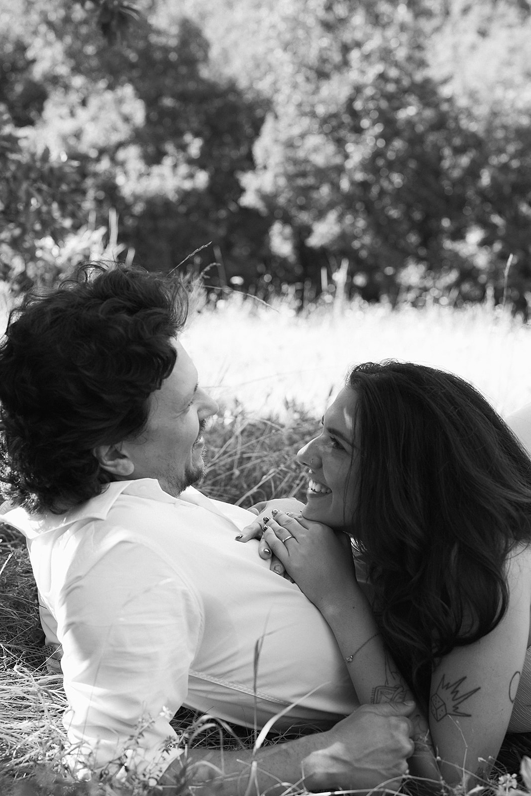 A couple dressed in white embraces outdoors, with greenery in the background during their candid engagement photos