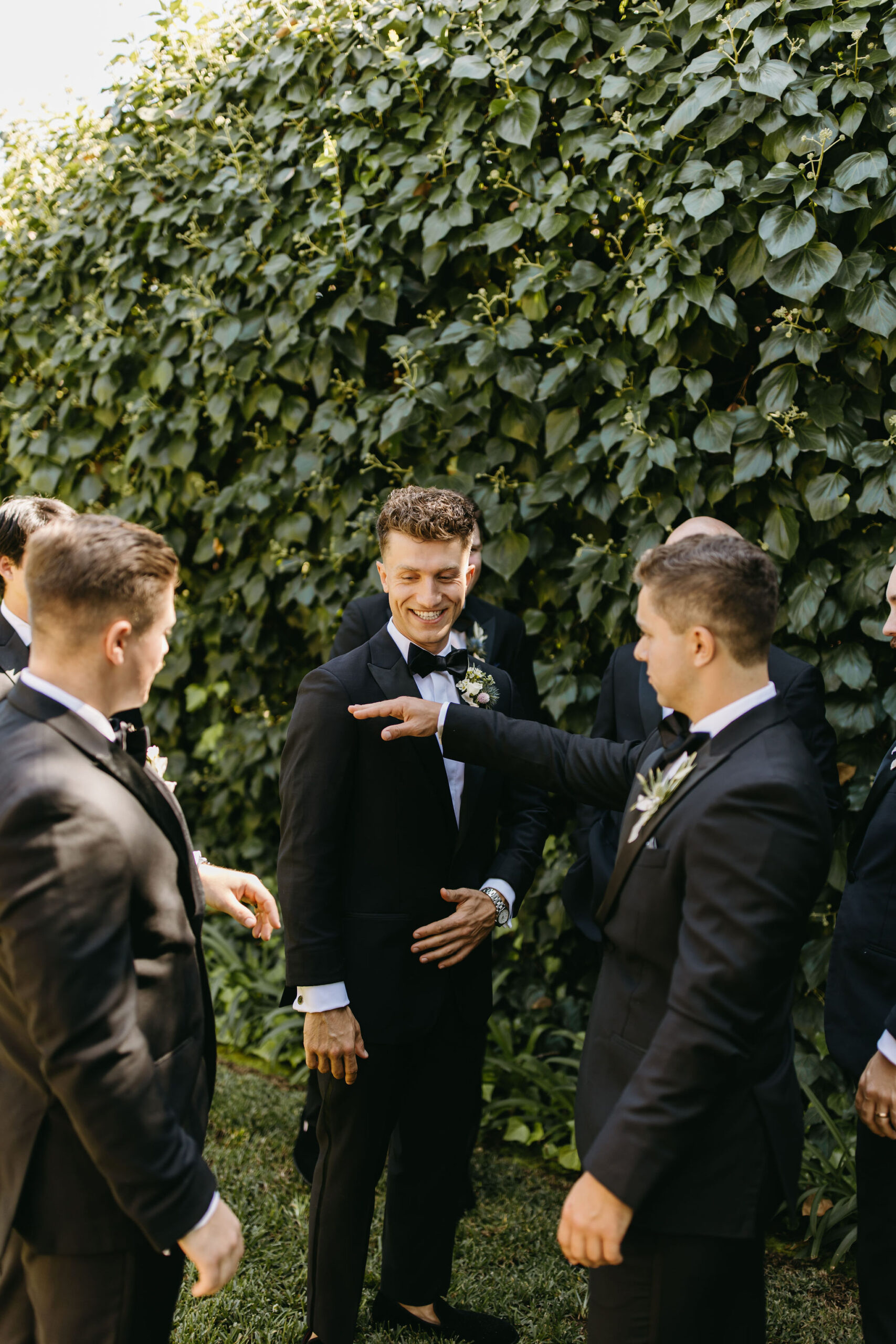 Group of men in suits standing and interacting outdoors with a green leafy background.