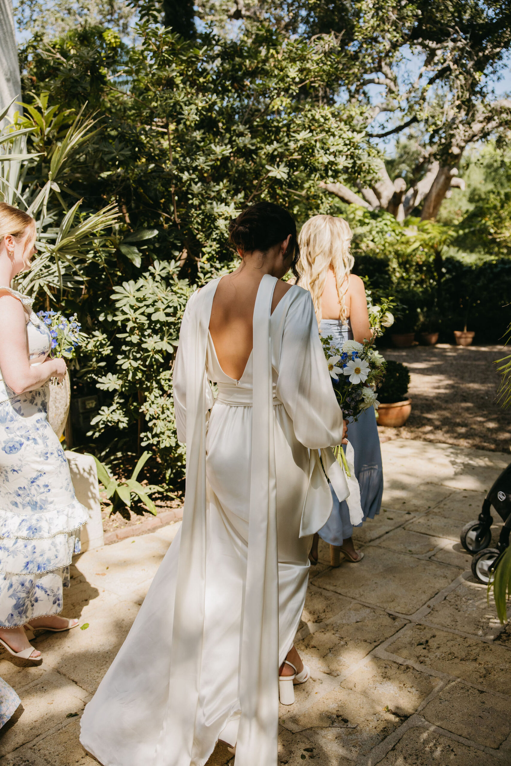 A woman in a white dress walks on a stone path surrounded by greenery, accompanied by others holding flowers.