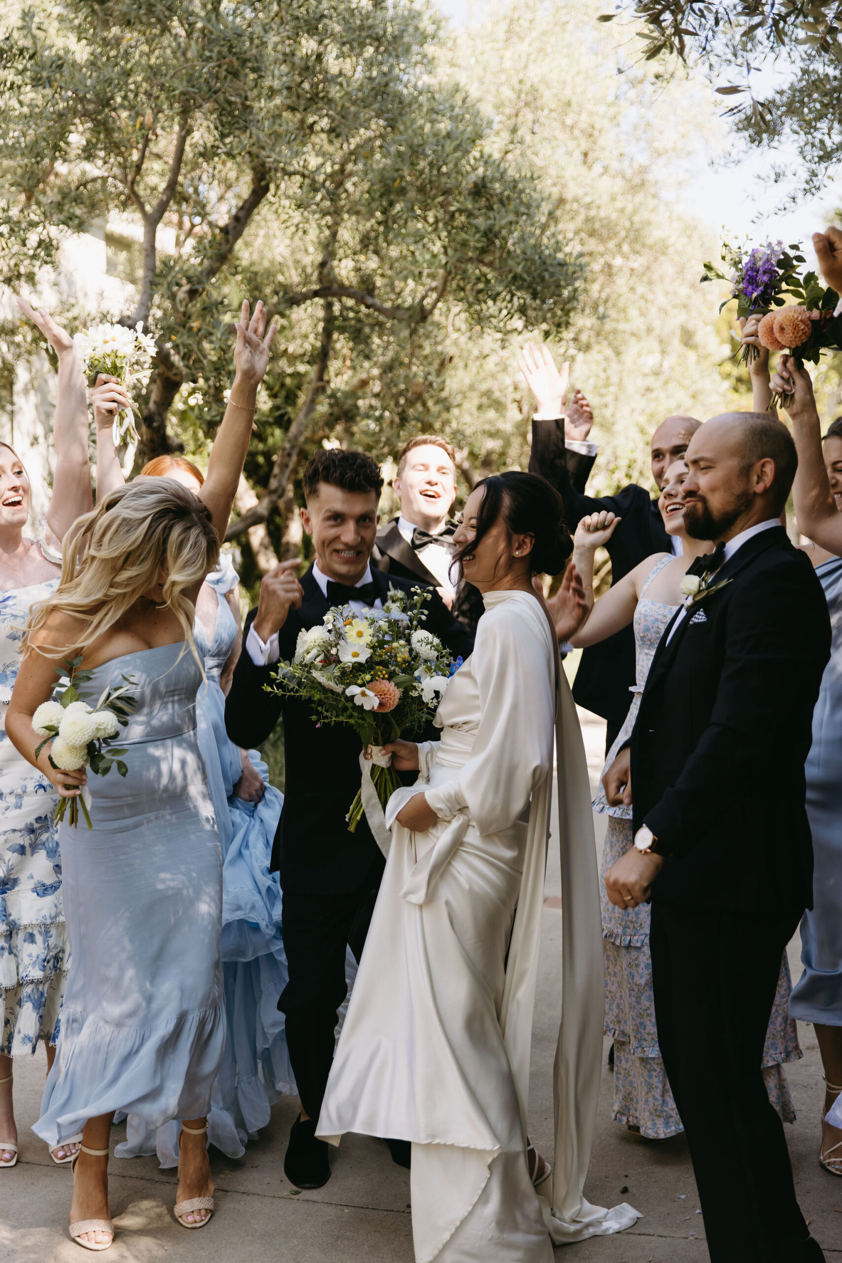 A bride and groom celebrate with a group of people outdoors. The bride holds a bouquet, and several attendees are cheering. Trees are visible in the background.