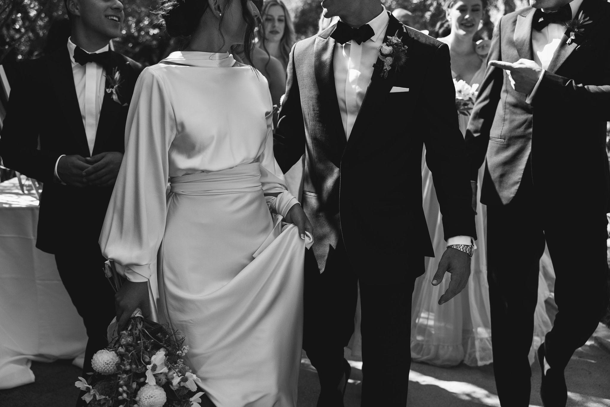 A bride and groom walk together, surrounded by their bridal party, all dressed in formal attire. The bride holds a bouquet. The scene is in black and white.