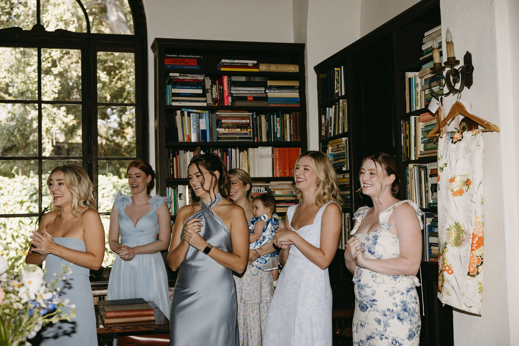 A group of six women in light dresses stand together, smiling, in a room with large windows and bookshelves.