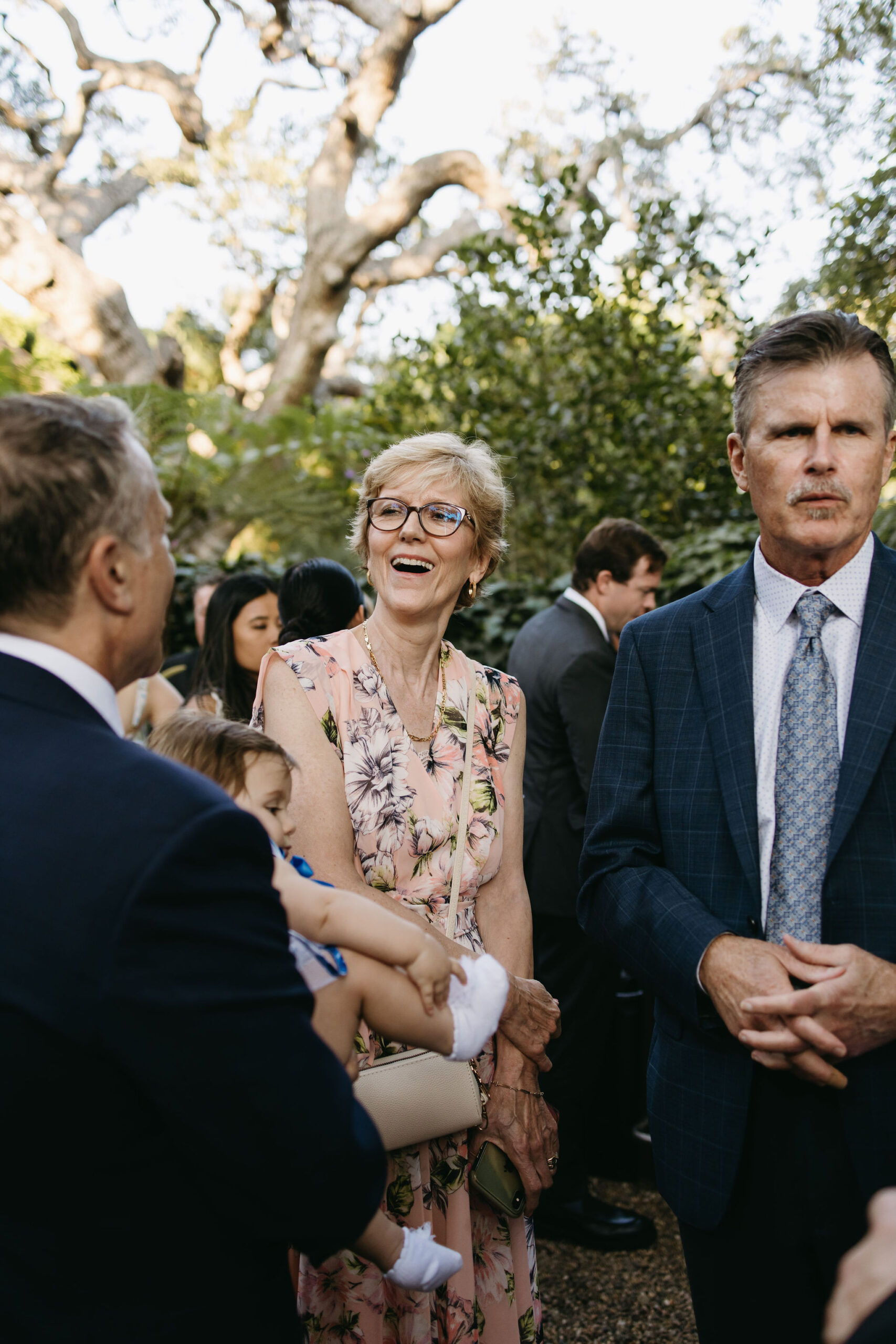 A group of people in formal attire gather outside at a garden event under a large tree.