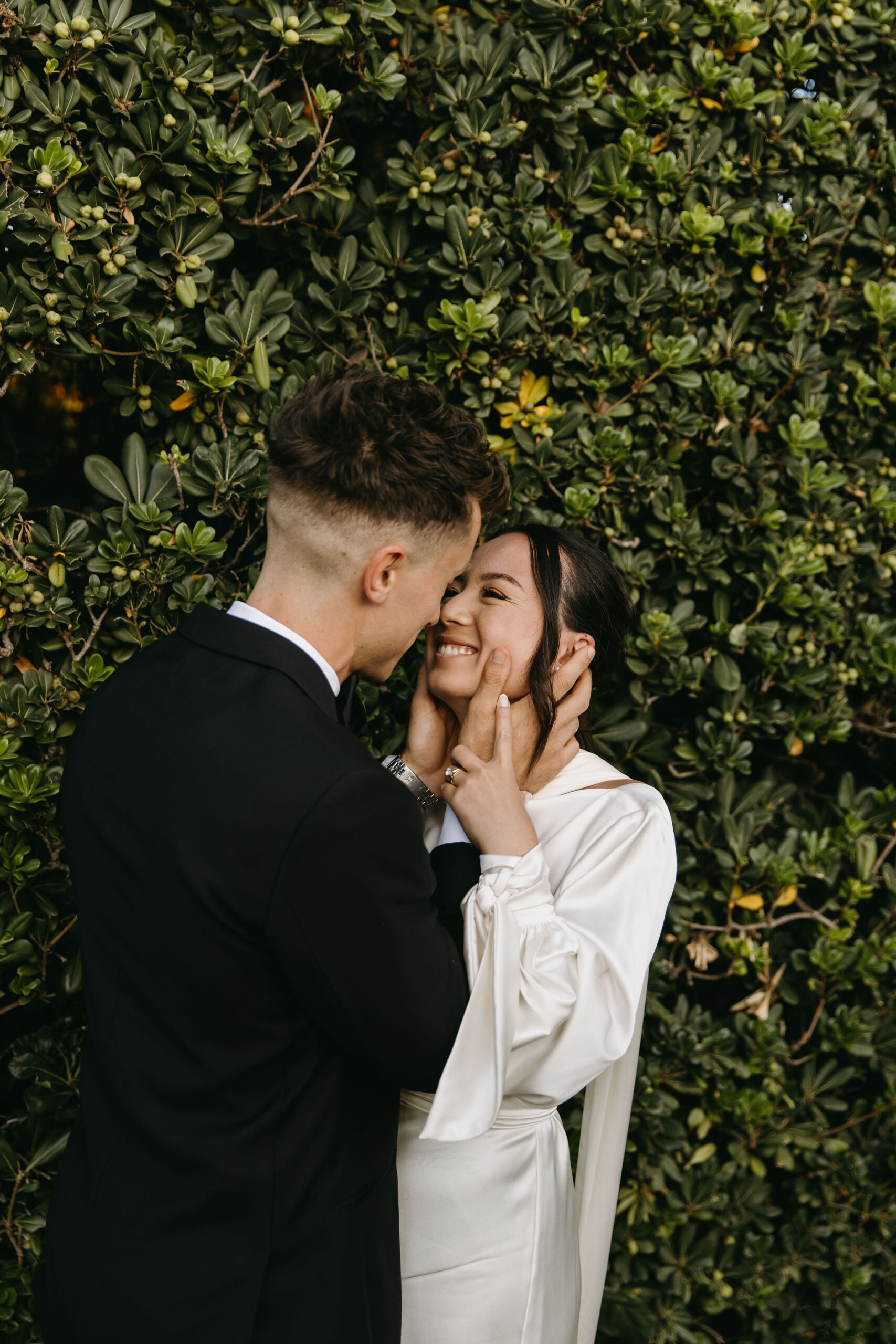 A couple in formal attire smiles and dances outdoors surrounded by greenery.