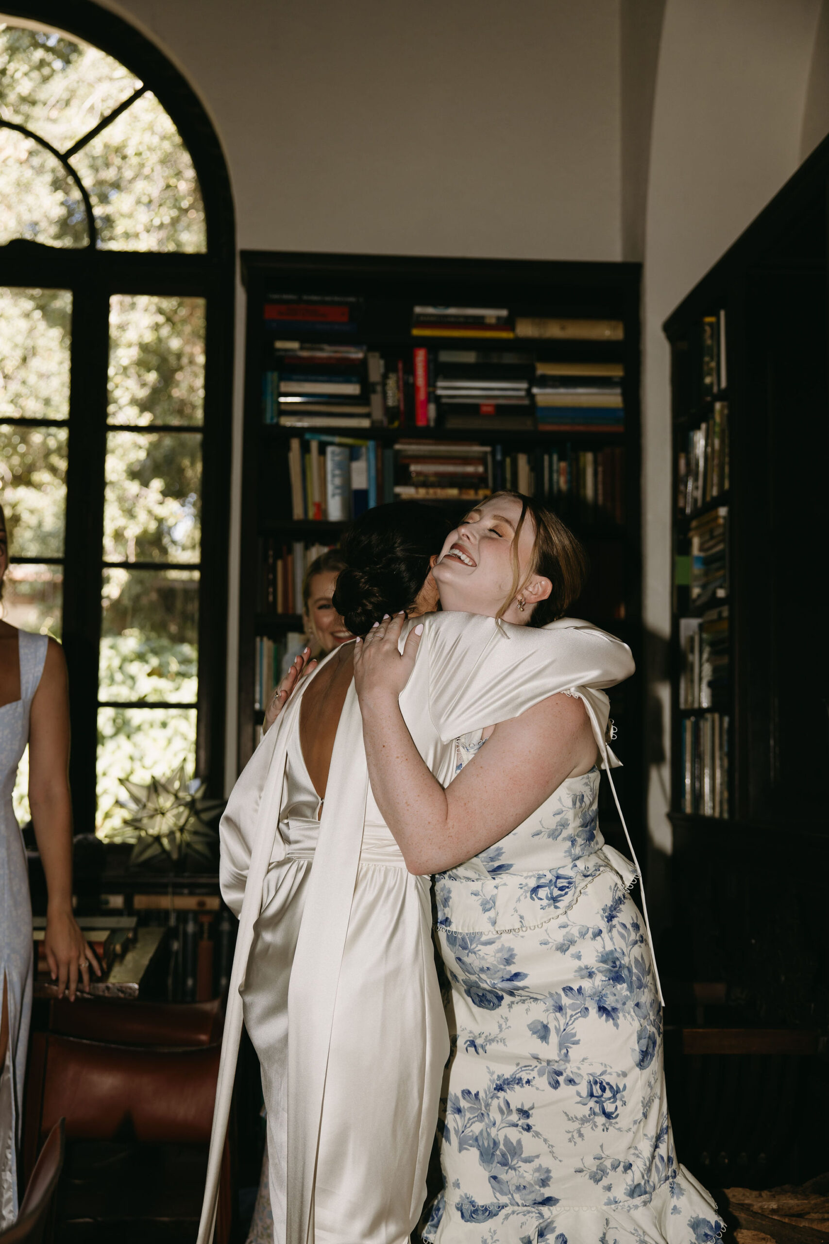 Two people embracing indoors, one in a white robe and the other in a floral dress, with bookshelves and a large window in the background.