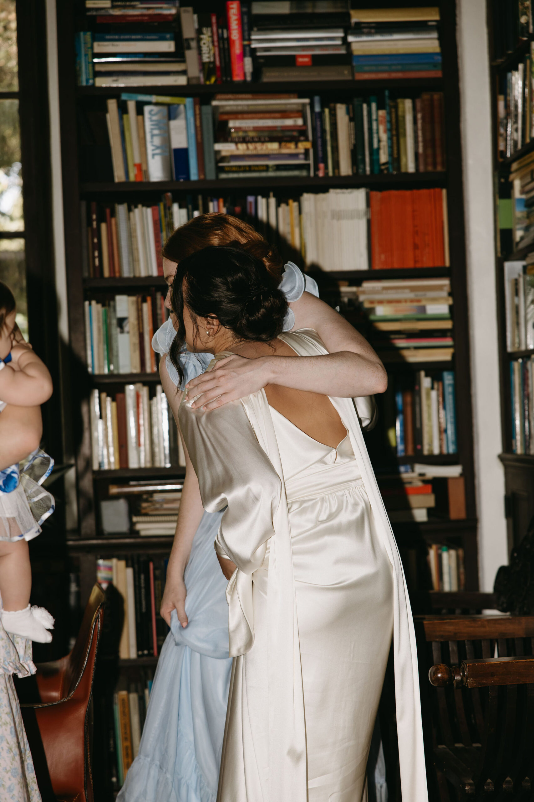 Two people embracing indoors, one in a white robe and the other in a floral dress, with bookshelves and a large window in the background.