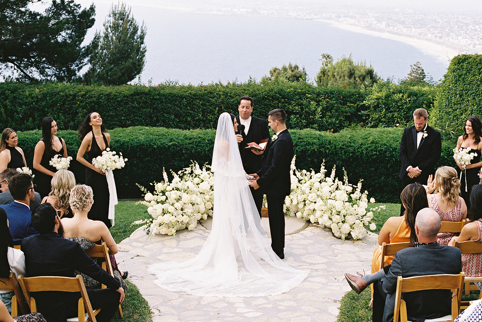 A couple is getting married outdoors, with an officiant and wedding party present. Guests are seated, and the background features lush greenery and a view of the sea.