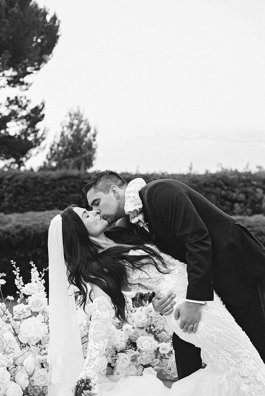 A couple stands before an officiant during an outdoor wedding ceremony with white flowers and a scenic background.