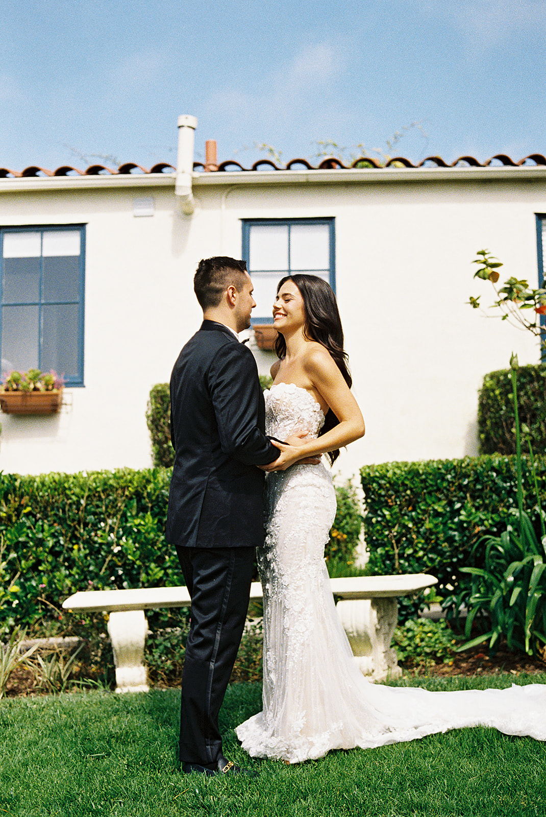 A bride in a white dress and a groom in a black suit stand facing each other in a garden, in front of a white house with blue-trimmed windows.