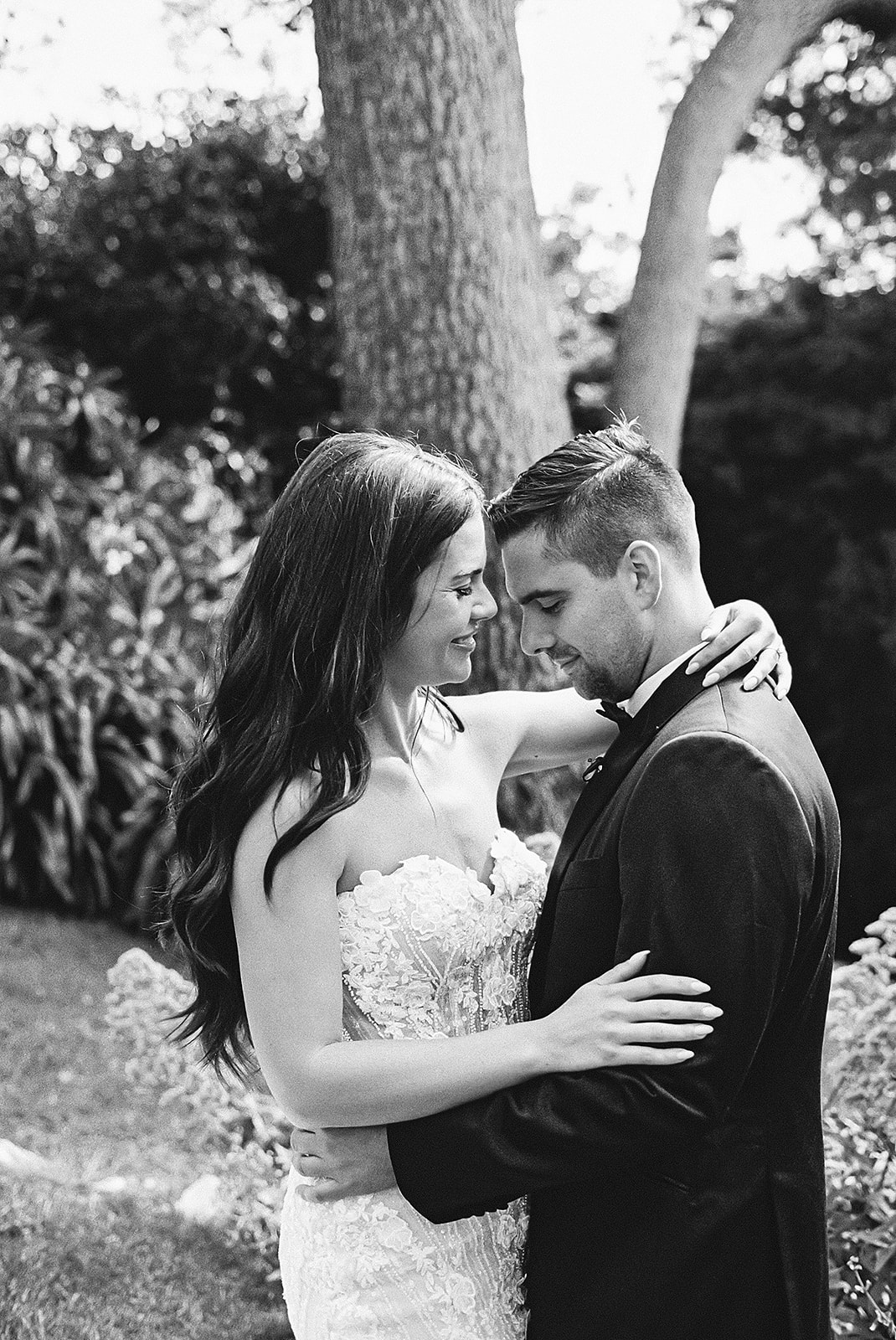 A bride and groom embrace outdoors, surrounded by greenery. The groom wears a suit, and the bride is in a strapless wedding dress.