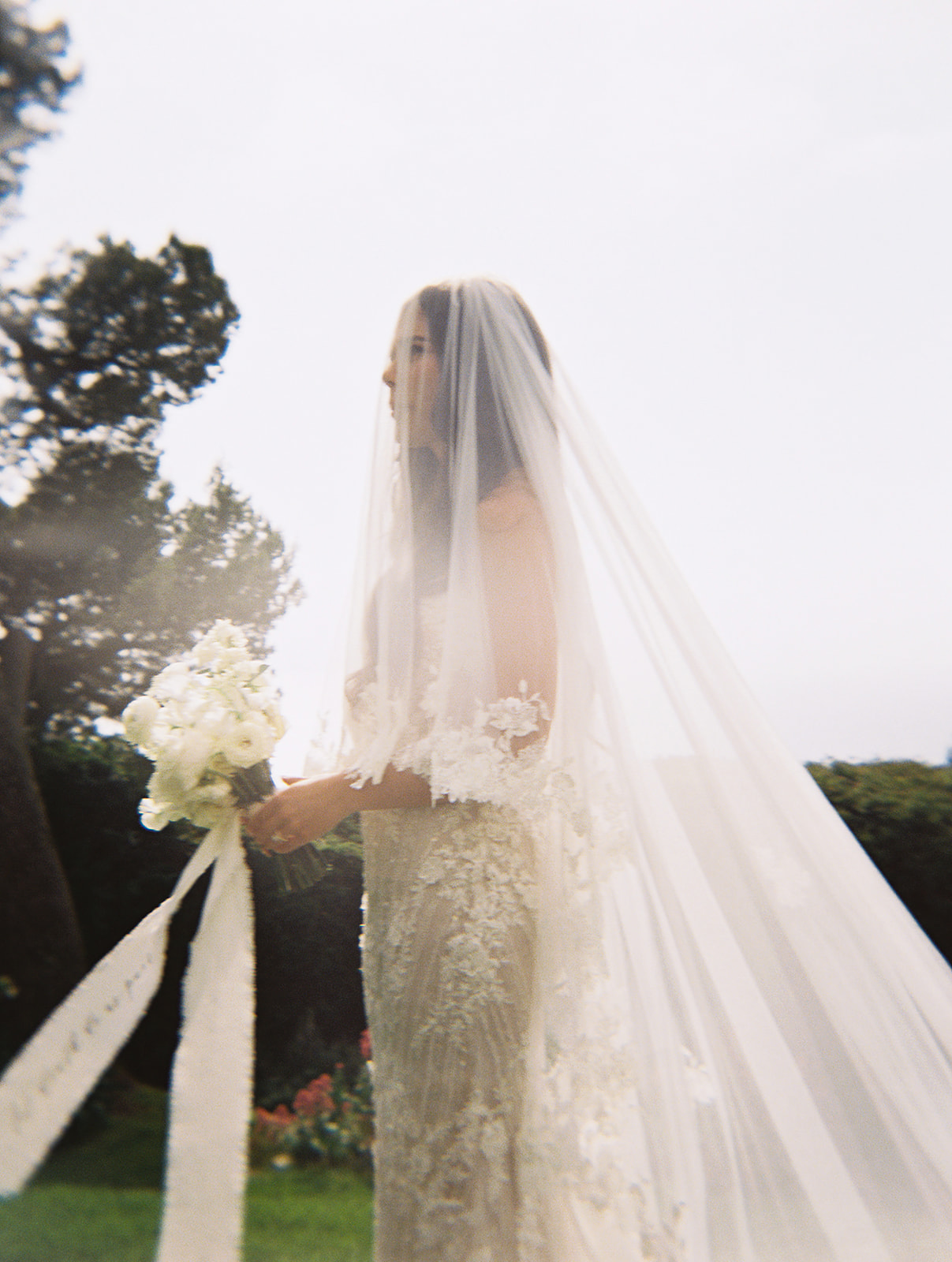 Bride wearing a white dress and veil holds a bouquet of white flowers, standing outdoors with greenery in the background.