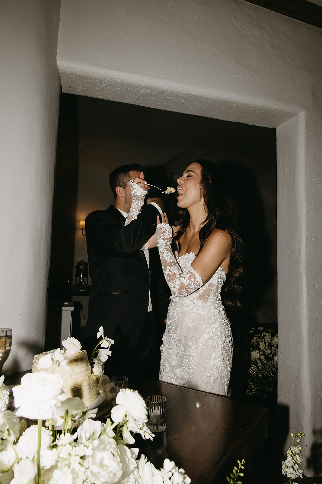 A bride and groom in formal attire cut a wedding cake together on a table adorned with white flowers and candles.