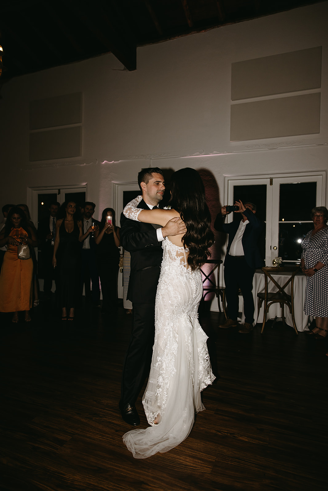 A bride and groom dance together in a dimly lit room, surrounded by guests.