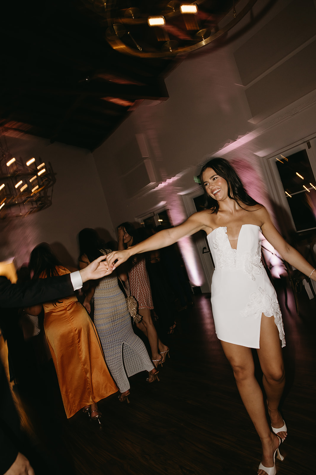 Woman in a white dress dancing with a man, amidst a group of people in formal attire at an indoor event.