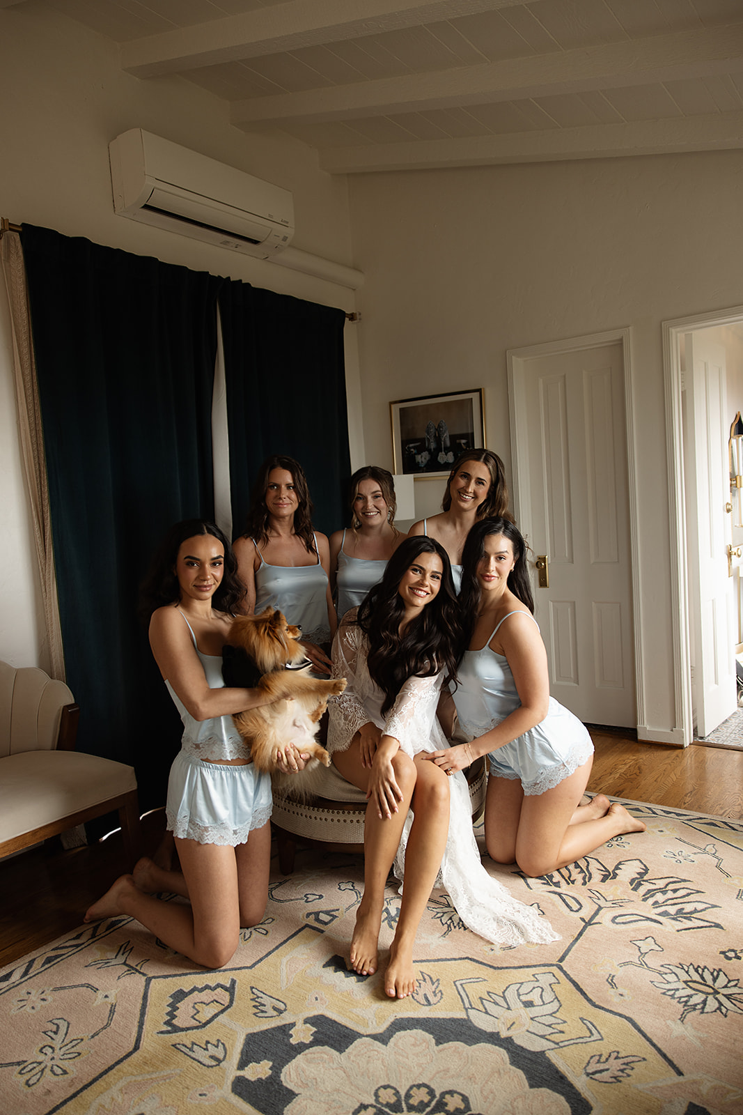 A group of bridesmaid in matching light blue attire pose indoors on a rug, with one holding a small dog for a wedding at the La Venta Inn