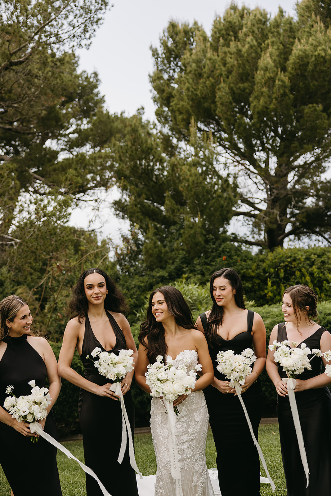 A bride stands with five bridesmaids, each holding white flower bouquets. The women are dressed in black dresses, and they are outdoors with trees and greenery in the background.