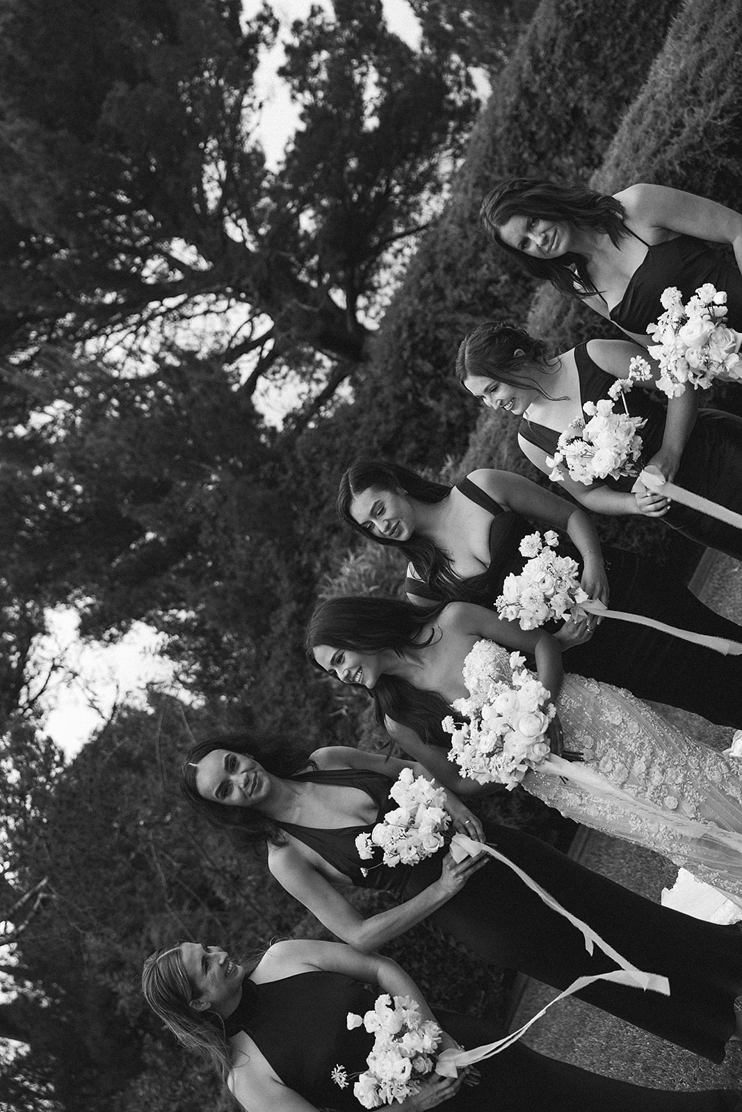 A bride stands with five bridesmaids, each holding white flower bouquets. The women are dressed in black dresses, and they are outdoors with trees and greenery in the background.