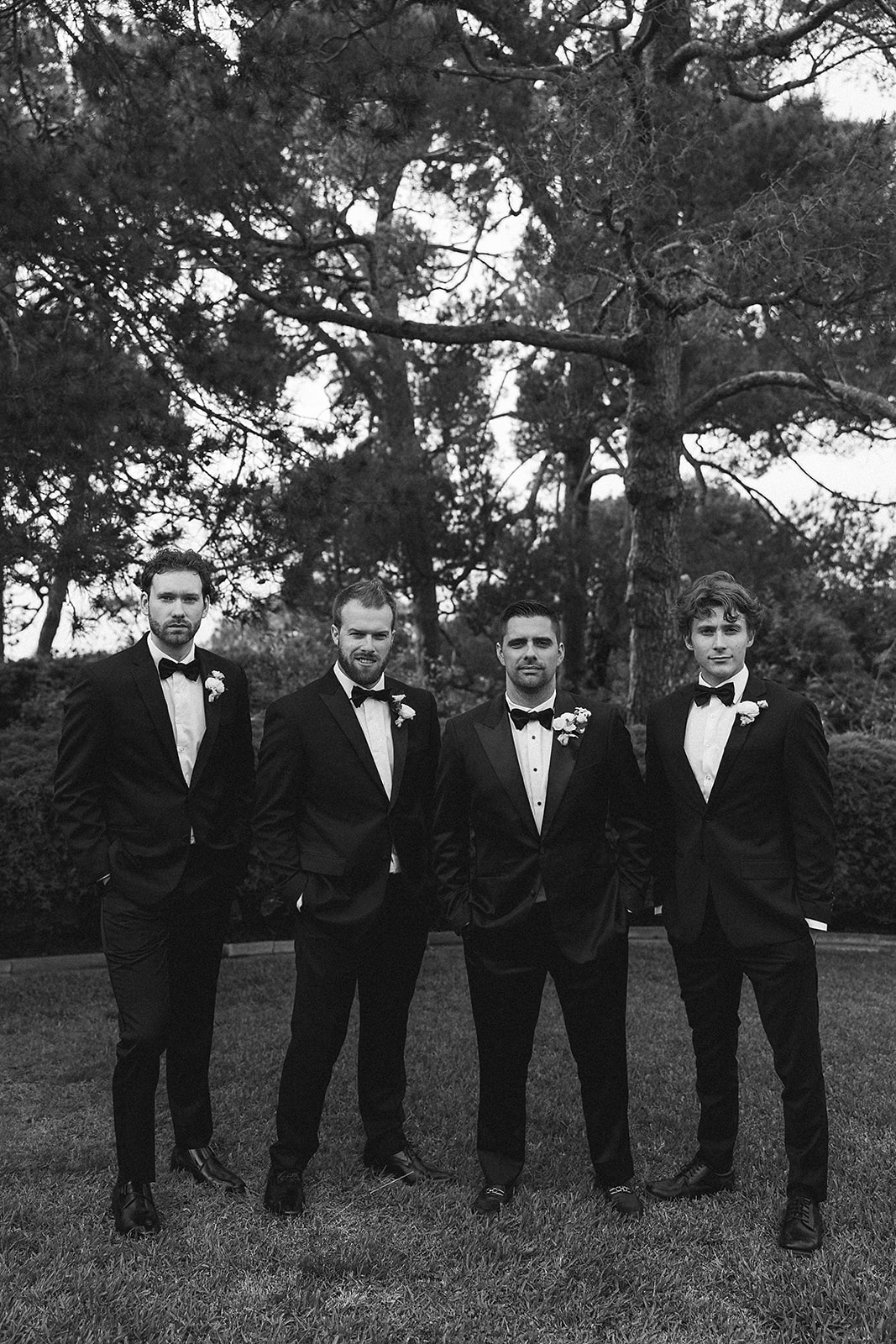 Four men in tuxedos with boutonnières stand outdoors, looking in different directions. Trees are visible in the background.