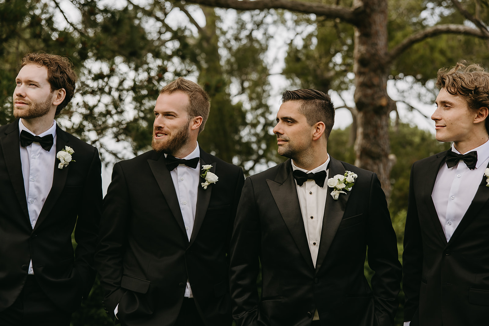 Four men in tuxedos with boutonnières stand outdoors, looking in different directions. Trees are visible in the background.