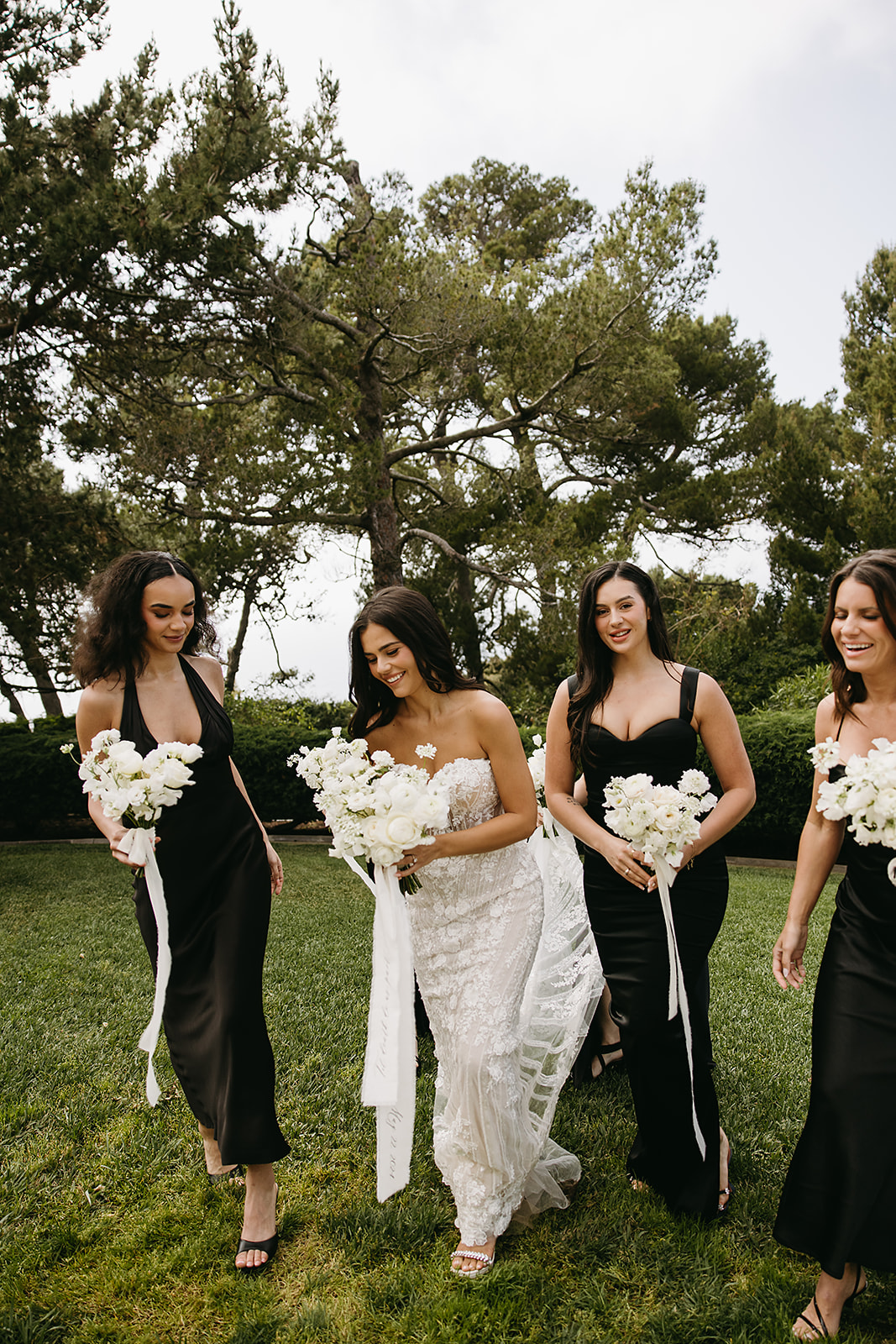 A bride stands with five bridesmaids, each holding white flower bouquets. The women are dressed in black dresses, and they are outdoors with trees and greenery in the background.